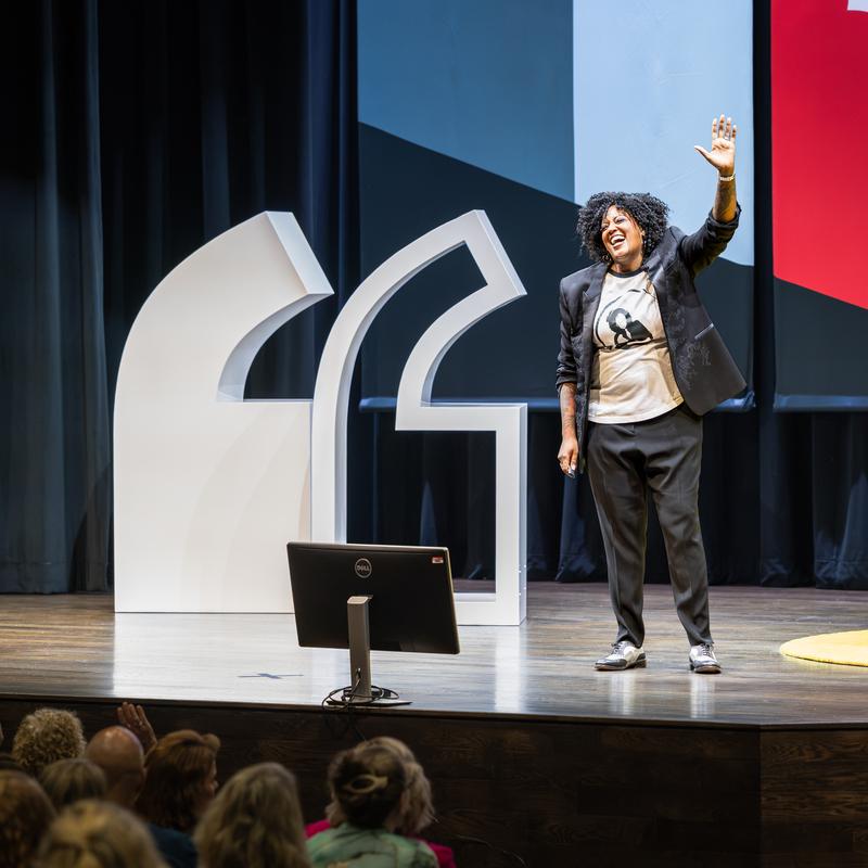 Cassandra Worthy speaks on stage at Aims Community College with a hand raised asking audience members to raise their hands